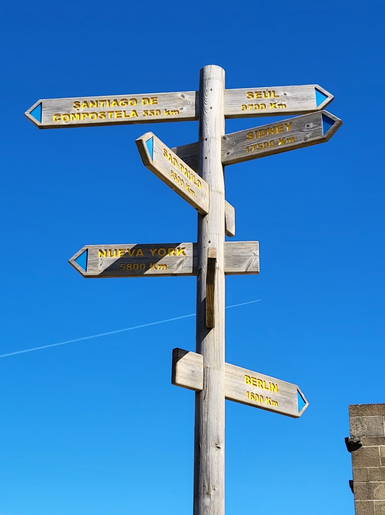 A wooden sign at the top of Alto de Perdón points to major cities around the world. A jet flies in the distant background of the beautiful blue sky.