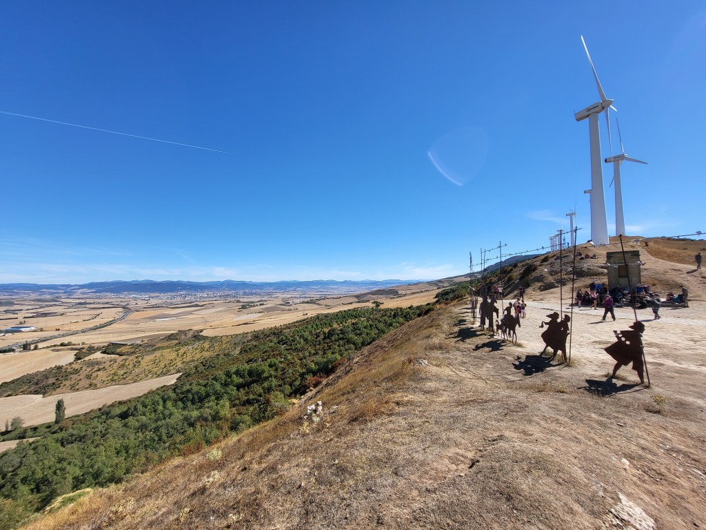 The metal statues at the top of the Alto de Perdón. Pilgrims rest between the statues and the windmills on top of the ridge. Pamplona is in the distant valley.