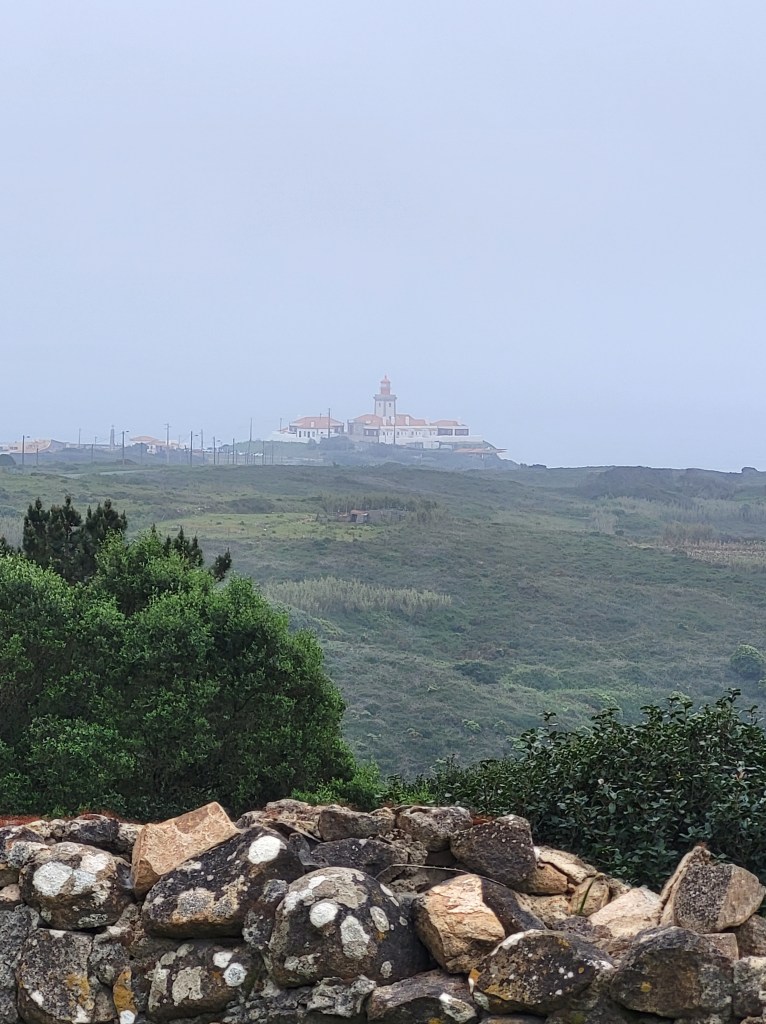 A lighthouse sits on a hill in the foggy distance. The hill is Cabo da Roca, the westernmost point of the European continent. The lighthouse is the Farol do Cabo da Roca.