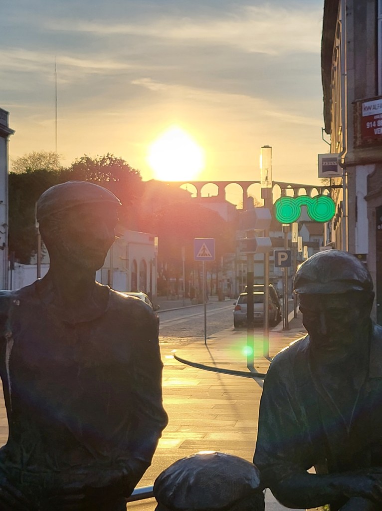 The sun rises in the background over the aqueduct. In the foreground, there is a statue of three men.