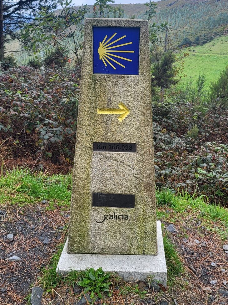 Signpost with a St. James shell, a yellow arrow, a distance marker, and inscribed with Galicia.